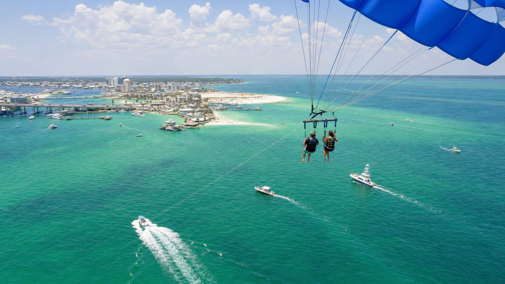 Couple parasailing high over the emerald green Destin Harbor with charter fishing boats below and the Gulf of Mexico stretching to the horizon