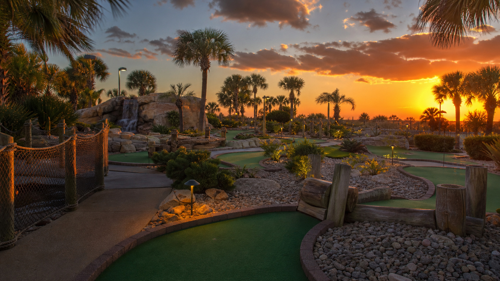 Late evening golden light over a tropical mini golf course in Destin Florida, course pathway lights switching on as the sun drops below the palm trees