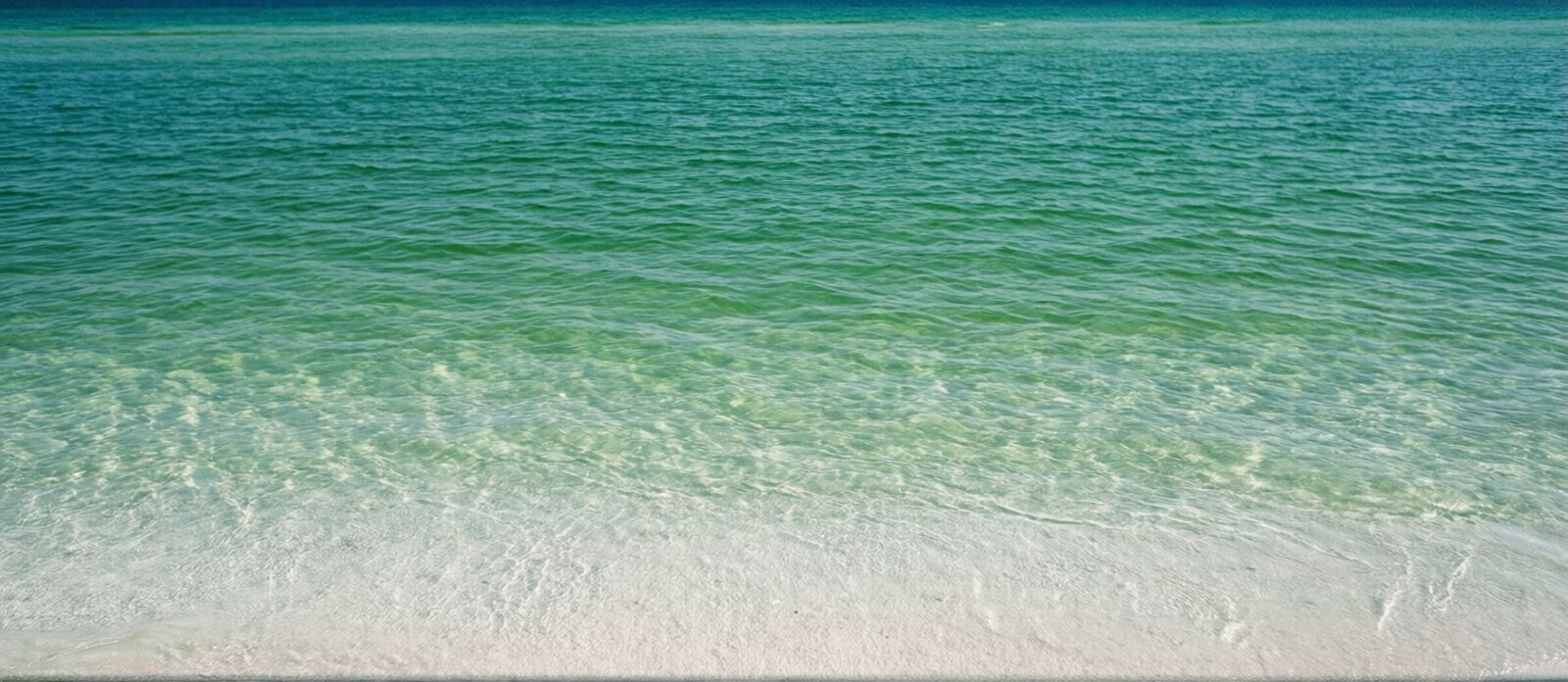 Crystal-clear emerald green Gulf of Mexico water seen from the shoreline at Destin Florida beach on a sunny summer day, white quartz sand visible beneath shallow turquoise water, gentle ripples